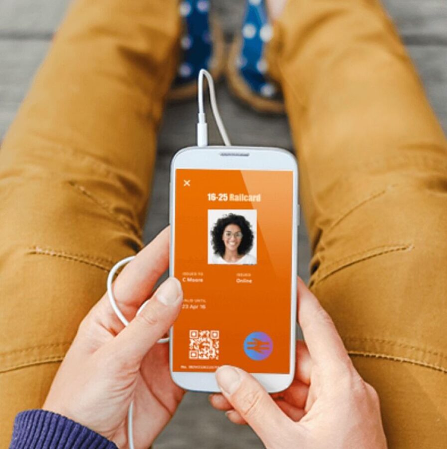 Pperson wearing brown pants holds a smartphone showing a digital 16-25 Railcard with a smiling individual’s photo.