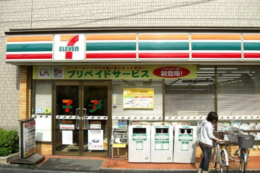 7-Eleven storefront with woman and bicycle, featuring recycling bins and greenery.