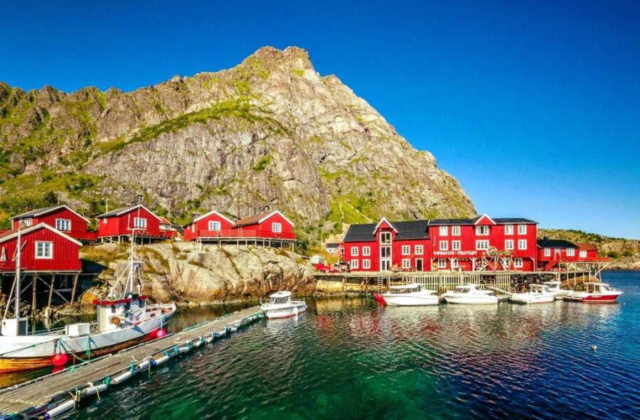 Å-Hamna Rorbuer red wooden cabins on stilts line a rocky shore in front of a towering cliff under a vivid blue sky. White boats float on the clear, greenish water.