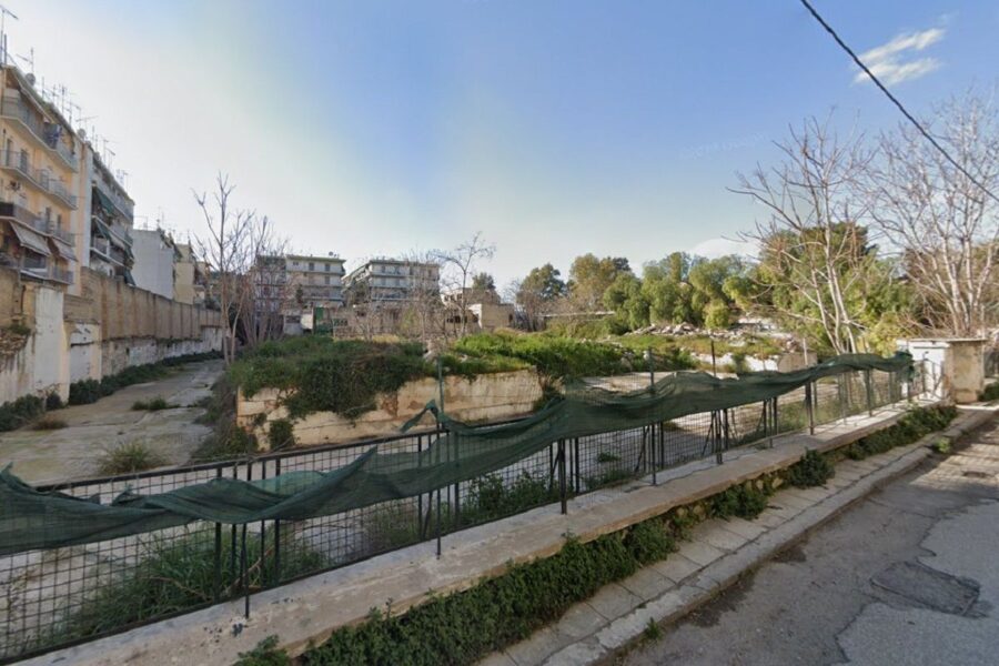 Abandoned overgrown urban lot with sunken terraces, concrete curb, railing, and adjacent apartment buildings.