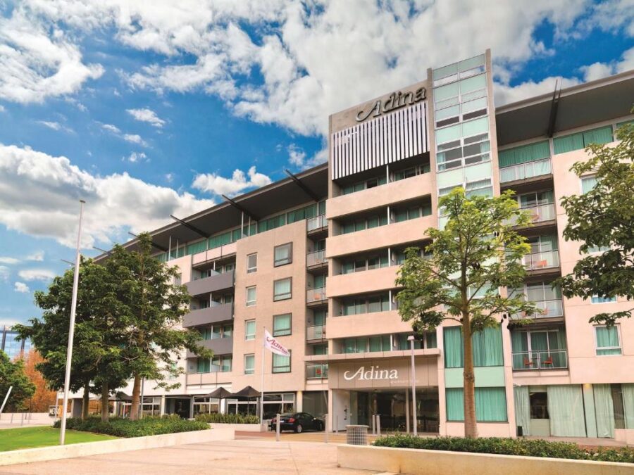 Adina Apartment Hotel Perth modern facade with balconies, trees and car under blue sky.