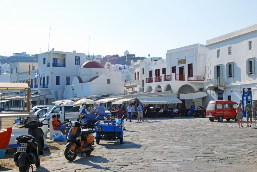 Bustling Aegean waterfront scene with scooters, whitewashed buildings, cafés, and blue skies.