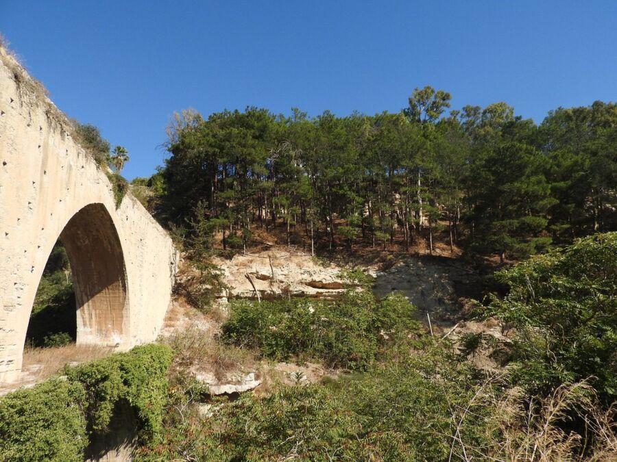 Scenic bridge spans a river in Agia Irini, Greece, surrounded by lush greenery and clear blue skies.