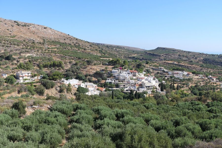 Agios Georgios village with white houses nestled on a hill, surrounded by lush olive groves and rugged terrain under a clear blue sky, conveying tranquility.