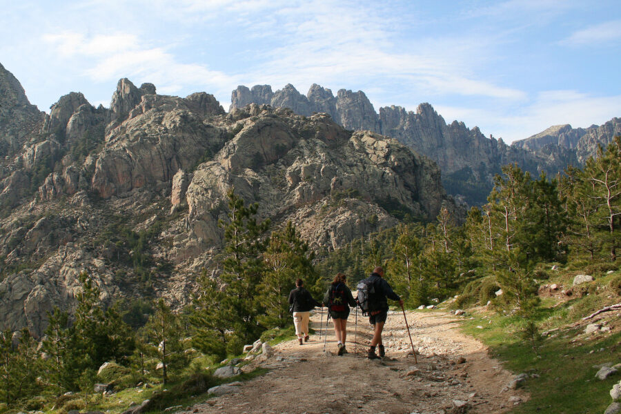 Several people hike along a rugged path in Aiguilles de Bavella, Corsica, with dramatic peaks in the background.