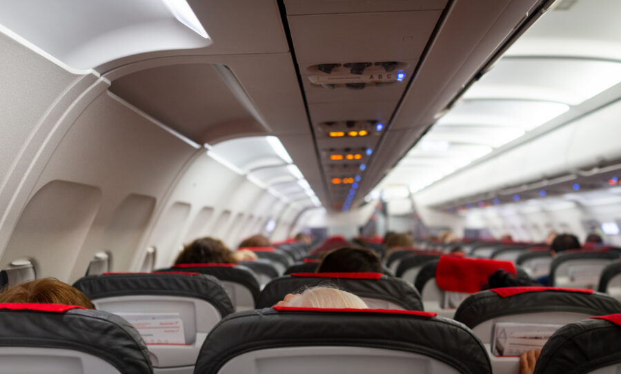 Airplane cabin interior with passengers, gray and red seats, overhead compartments, and soft lighting.