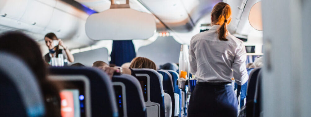Interior of an airplane with passengers seated and a stewardess in uniform serving drinks in the aisle during a flight.
