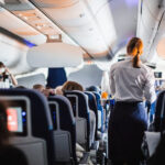 Interior of an airplane with passengers seated and a stewardess in uniform serving drinks in the aisle during a flight.