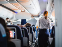Interior of an airplane with passengers seated and a stewardess in uniform serving drinks in the aisle during a flight.
