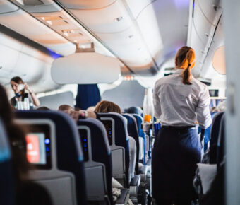 Interior of an airplane with passengers seated and a stewardess in uniform serving drinks in the aisle during a flight.