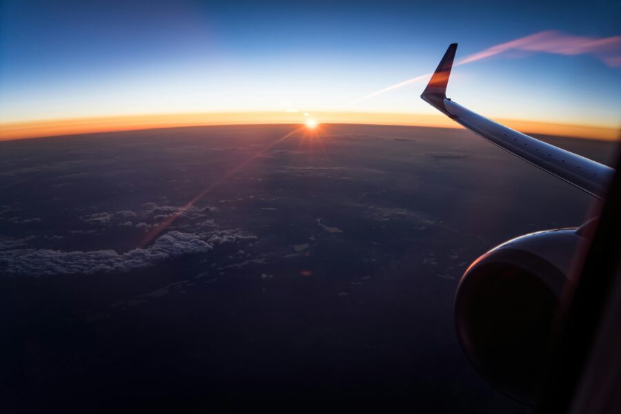 Airplane wing silhouetted against sunrise horizon, aerial travel view from window seat.
