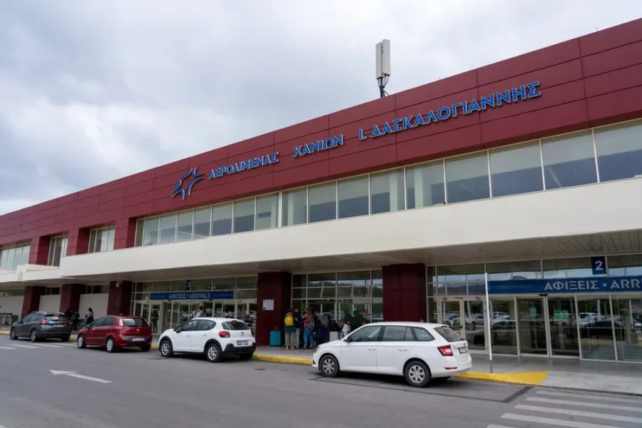 Airport arrivals terminal exterior with travelers, parked cars, and bilingual Greek-English signage.