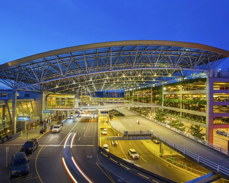 Portland airport arrivals terminal at twilight with curved canopy, parking garage, and traffic light trails.