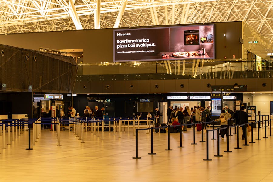 Modern airport concourse with check-in queues, stanchions, travelers, and illuminated Hisense pizza advertisement.