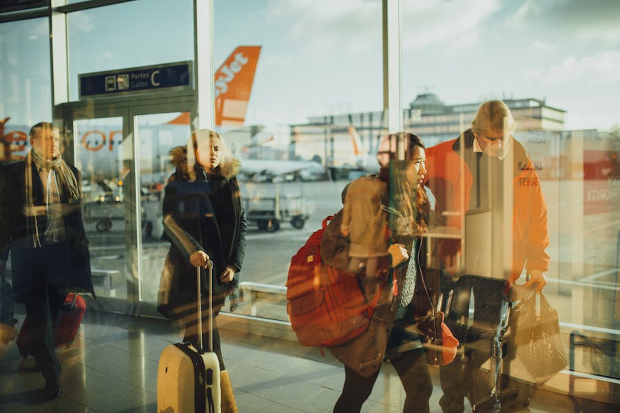 Airport terminal with travelers, luggage, glass reflections and orange airplane tail at boarding gates.