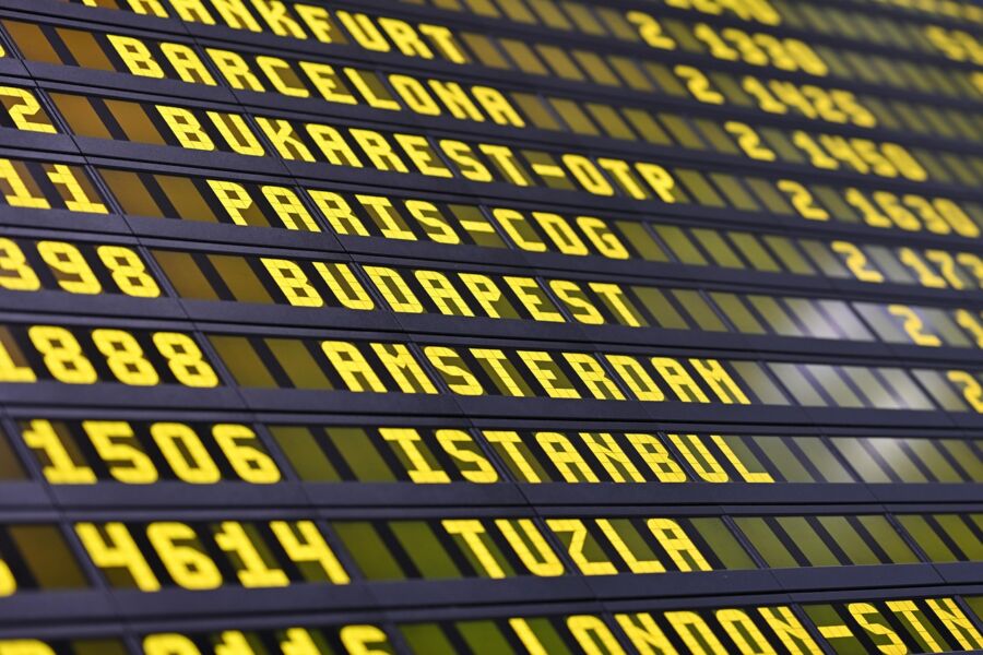 Airport digital flight board displaying European destinations like Frankfurt, Barcelona, Amsterdam, with flight numbers.