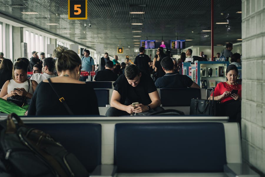 Airport gate 5 waiting area with seated travelers, luggage, phones, departure screens.
