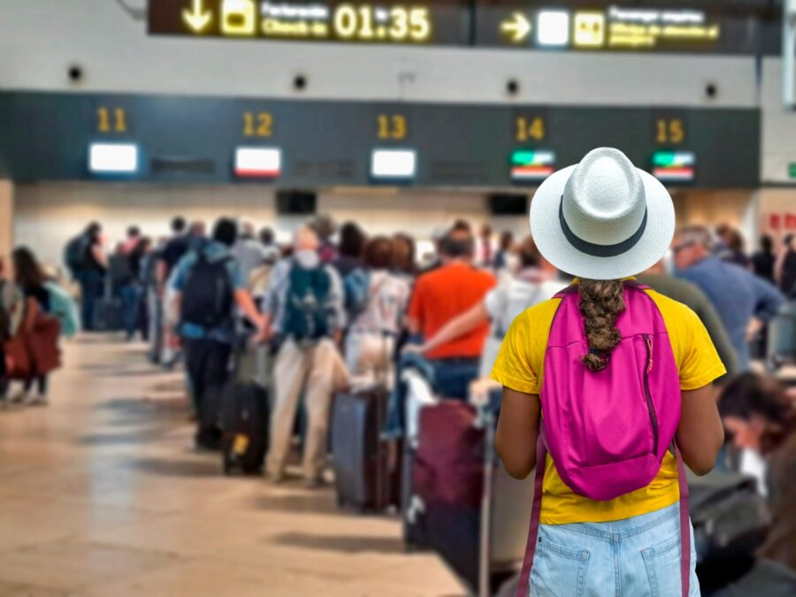 Young tourist stands in a long line at a busy airport check-in counter, surrounded by other travelers.