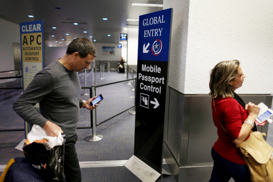 Travelers in airport terminal using phones, signs for Global Entry and Mobile Passport Control.