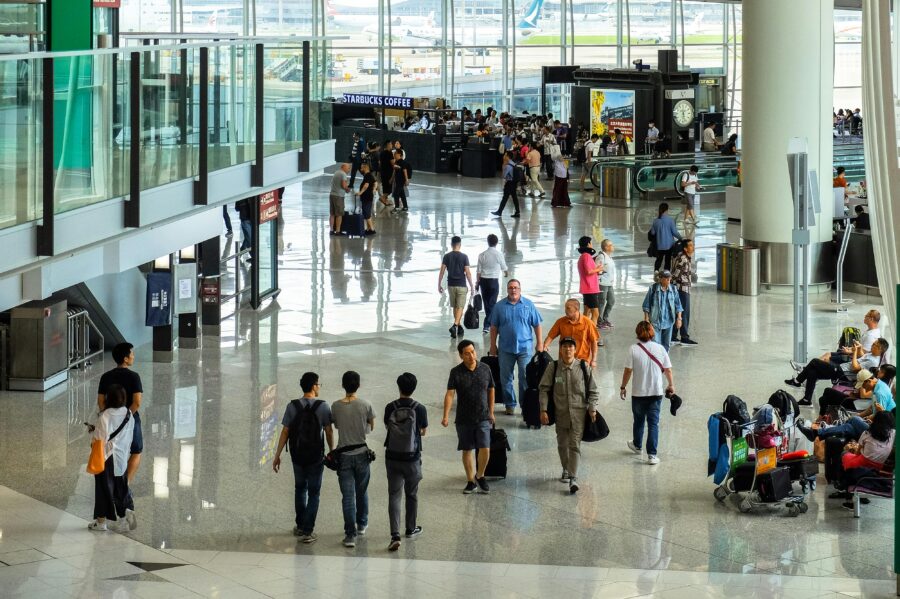 Bustling airport terminal with diverse travelers, luggage, Starbucks, and planes visible through windows.
