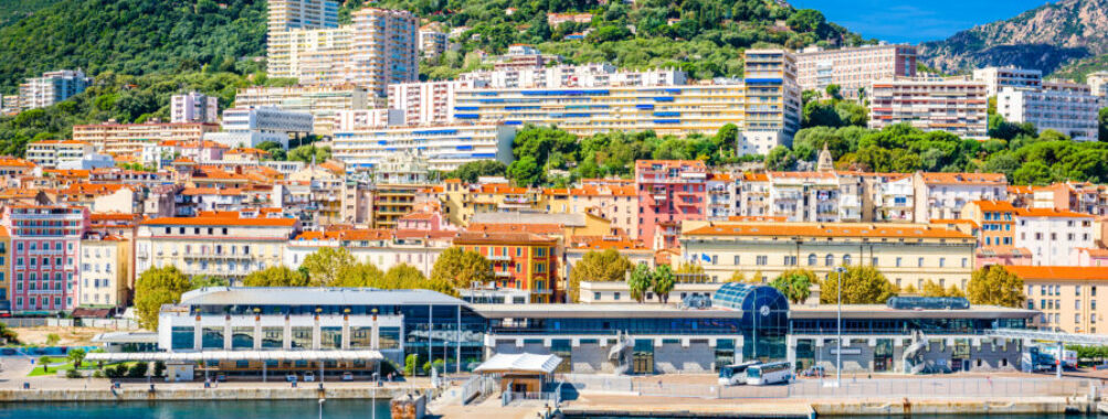 Coastal skyline of Ajaccio, Corsica, featuring colorful buildings and the Mediterranean Sea under a clear blue sky.