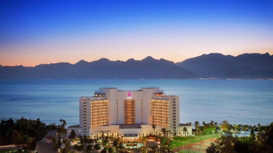Akra Hotel seaside resort at sunset with illuminated building, palm trees and mountain coastline view.