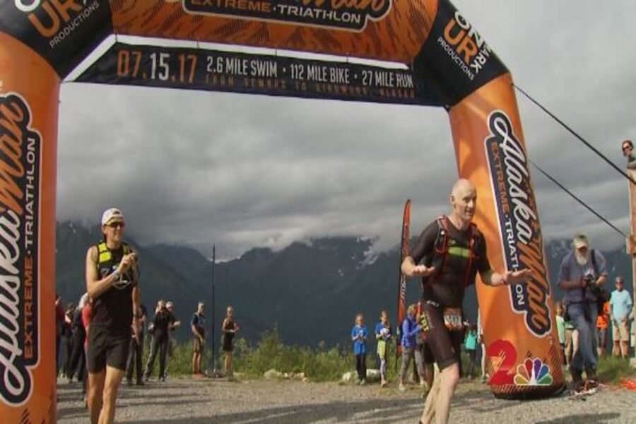 Athletes finish AlaskaMan Triathlon under arch, with cheering crowd and Alaskan landscape backdrop.