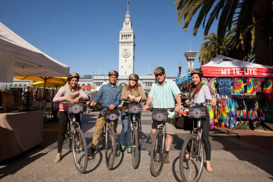 Group of five people on bicycles pose in front of a tower with a clock on Alcatraz Tickets and San Francisco's Streets Bike Tour. They wear helmets, and stalls selling colorful clothing are on the right.