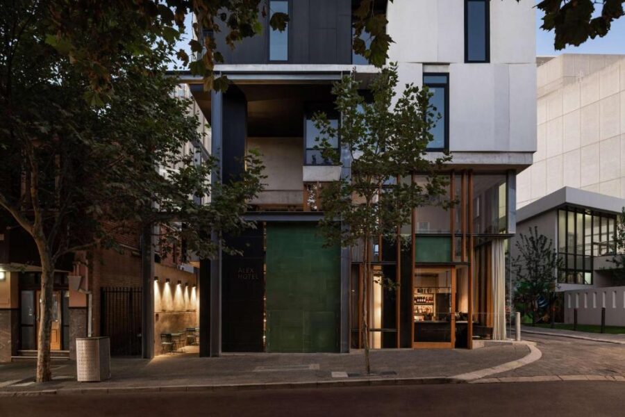 Modern Alex Hotel facade at dusk with glass entrance, balcony, and tree-lined urban street.