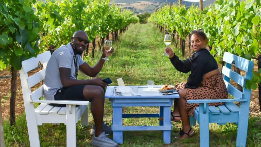 Smiling couple toasts with wine in a lush vineyard during a Alghero Wine Tasting. They sit on colorful benches, with a small table between them, conveying a joyful, relaxed ambiance.
