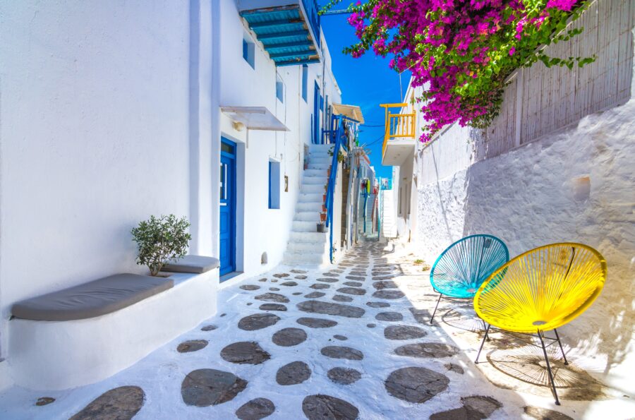 Charming Mediterranean alleyway with blue doors, vibrant bougainvillea, modern chairs, and stone path.