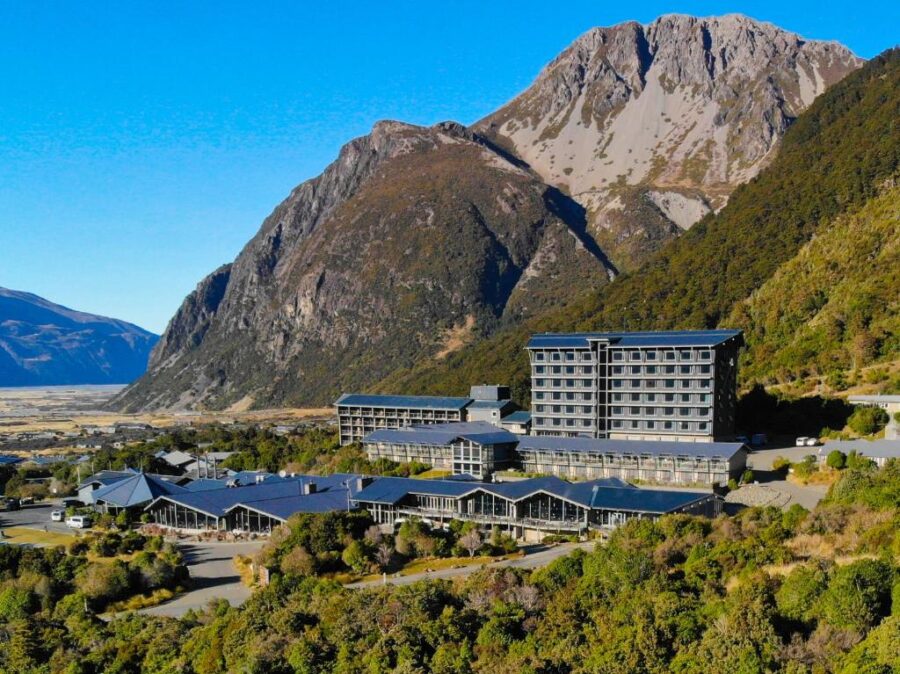 Luxury alpine mountain resort hotel beneath rugged peaks in sunny valley landscape.