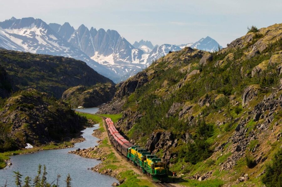White Pass & Yukon Route freight train along alpine lakeside amid rugged mountain peaks.