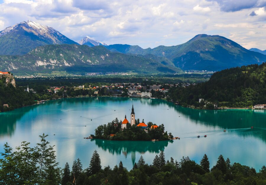 Aerial view of turquoise alpine lake with island church and surrounding mountains.