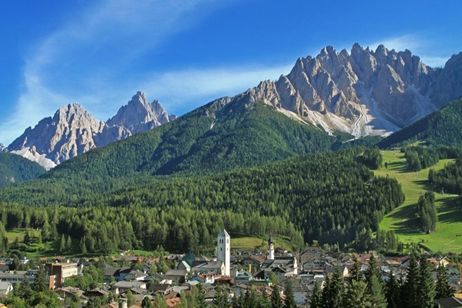 Alpine village with church tower under rocky mountain peaks and forested slopes.
