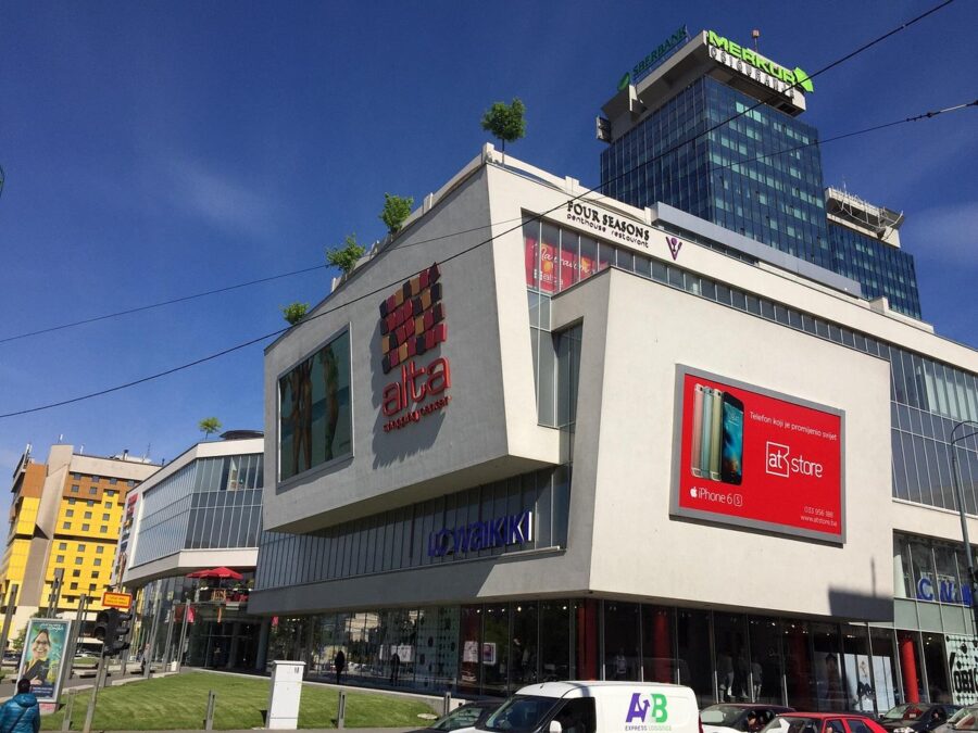 Alta Shopping Center features modern architecture, vibrant ads, and urban greenery under a clear sky.