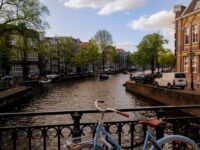 Amsterdam canal scene with a blue bike, lush trees, iconic architecture, and boats.