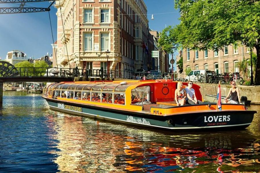 Amsterdam canal cruise: orange boat on sunny day, historic architecture, bridge, bicycles, lush greenery.