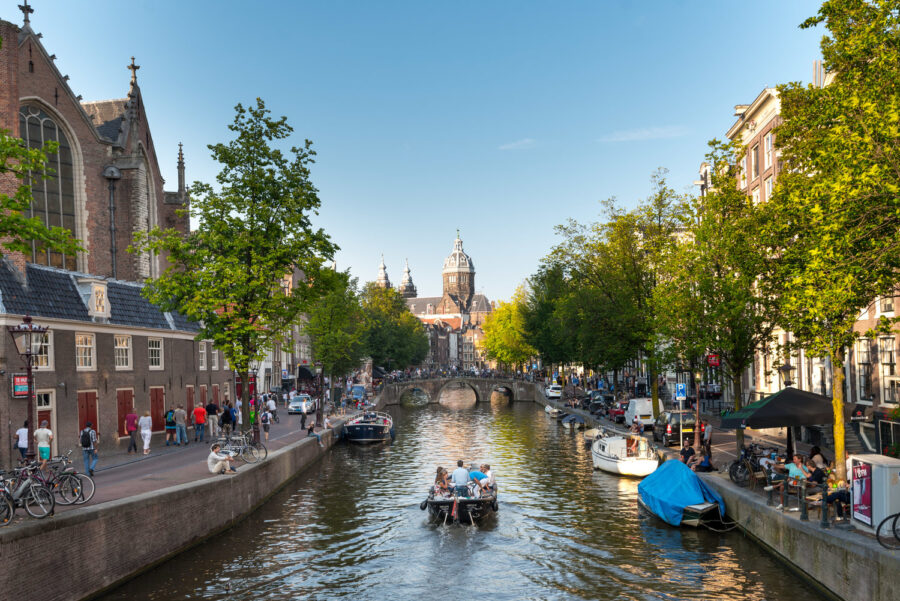 Amsterdam canal scene with boat, historic church, trees, pedestrians, and classic architecture.