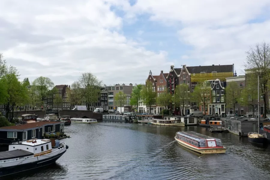 Amsterdam canal with tour boat, houseboats, historic gabled buildings, and spring trees under cloudy sky.