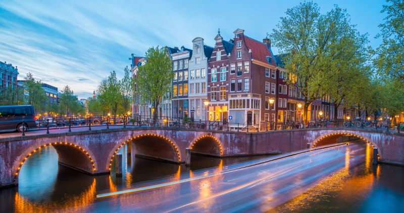 Amsterdam canal at twilight with lit bridge, historic townhouses, and reflections.
