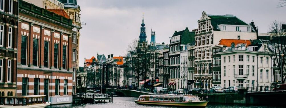 Amsterdam canal scene with historic architecture, boats, and overcast sky.
