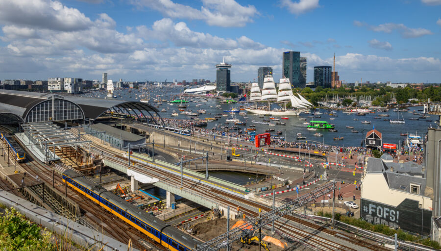 Amsterdam Central Station filled with NS trains and trams, surrounded by a vibrant crowd along the IJ river.