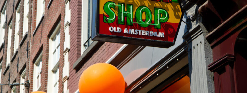Street view of an Amsterdam coffeeshop sign with vibrant red and green colors. Orange balloons hang nearby, adding a festive atmosphere