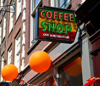 Street view of an Amsterdam coffeeshop sign with vibrant red and green colors. Orange balloons hang nearby, adding a festive atmosphere