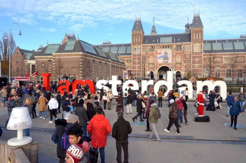 Tourists at I Amsterdam sign with Rijksmuseum backdrop on a sunny day.