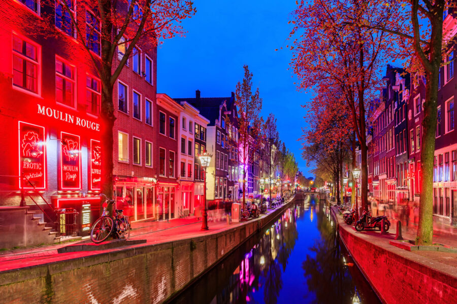 Amsterdam Red Light District canal at night with neon lights, reflections, bicycles, and historic buildings.