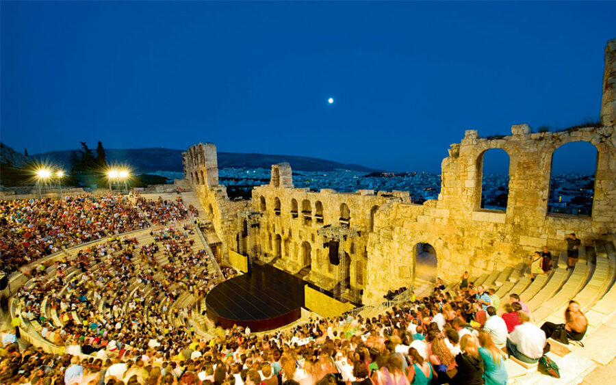 Ancient Greek amphitheater at twilight during live performance with audience and city backdrop.