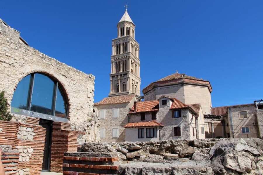 Mediterranean town with historic bell tower, ancient ruins, stone houses, and blue sky.
