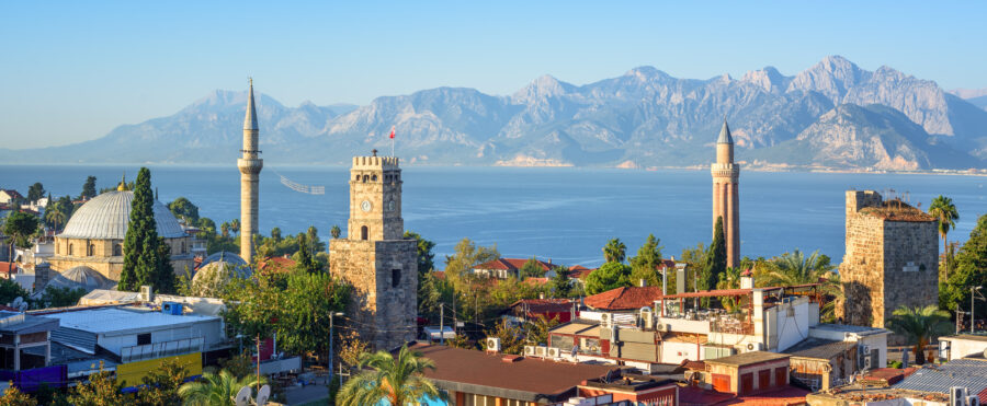 Scenic view of Antalya, Turkey, featuring historic towers, domes, and lush greenery against the backdrop of a calm sea and rugged mountains under a clear sky.